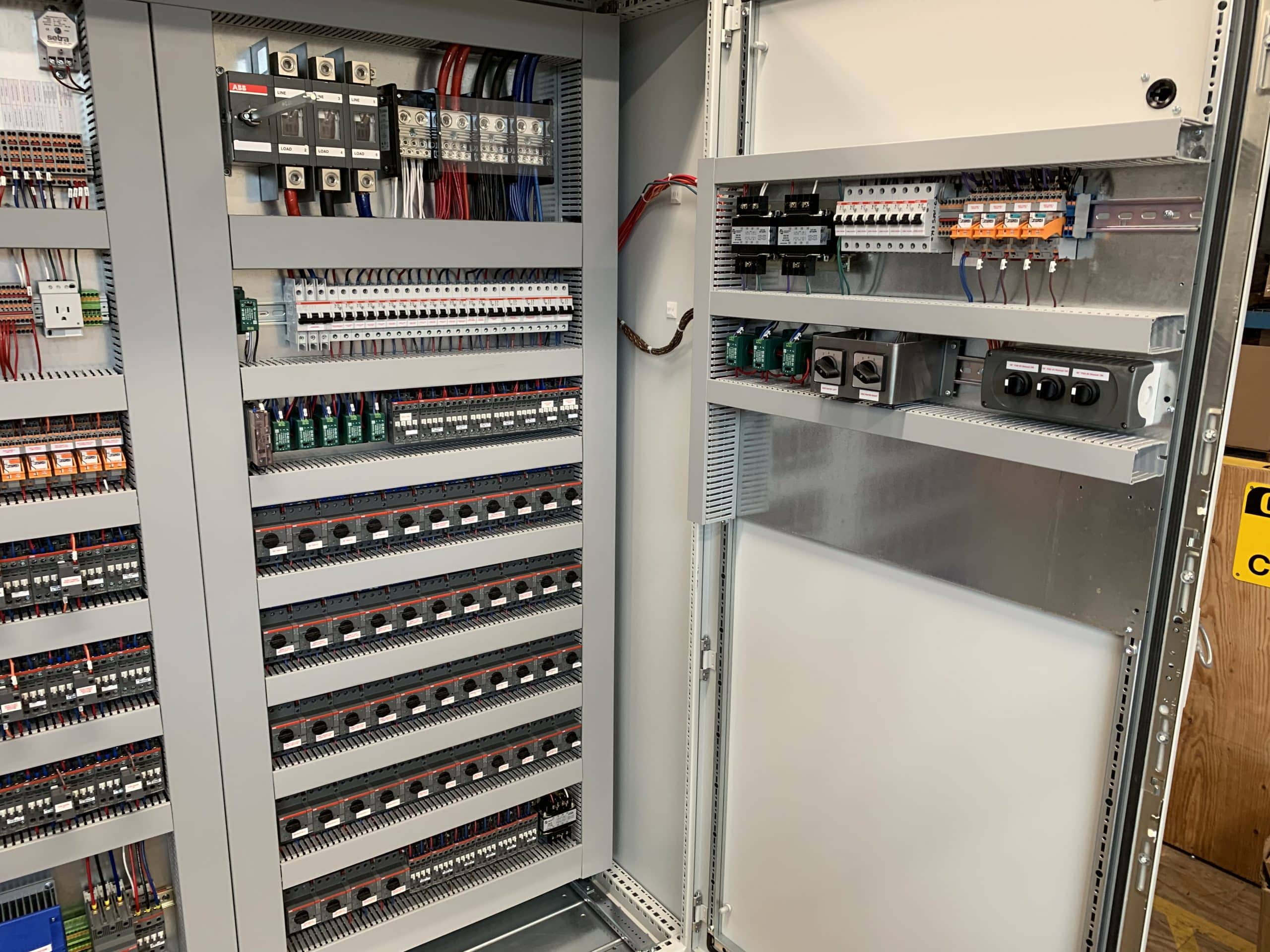 Open industrial electrical control panel with circuit breakers, terminal blocks, relays, and neatly arranged wiring.