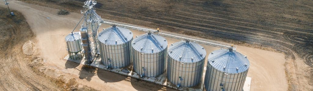 Aerial view of six large cylindrical grain silos with conical roofs, connected by walkways in an agricultural field.